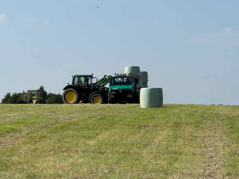 John Deere Traktor mit Frontlader belädt Unimog mit angehängtem Ballenwagen