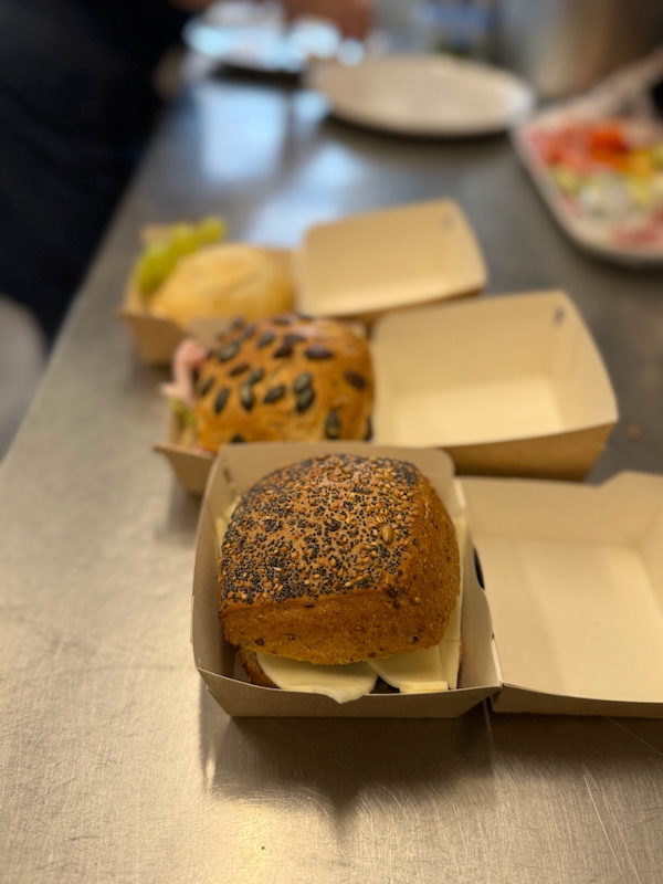 Drei belegte Brötchen zum Mitnehmen in Papp-Faltschachteln auf Arbeitsfläche der Hofküche.