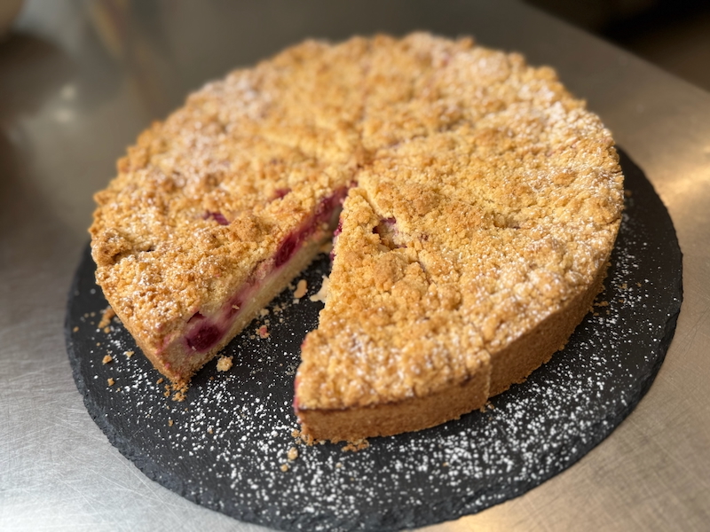 Angeschnittener Himbeer-Pudding-Streusel, leicht mit Puderzucker bestreut auf runder Schieferplatte.
