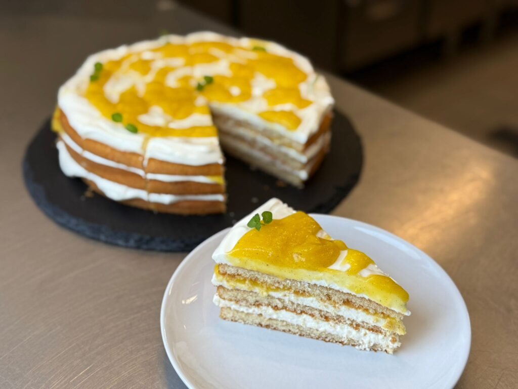 Sahnetorte mit Mango angerichtet auf weißem Porzellanteller und der Tort im Hintergrund