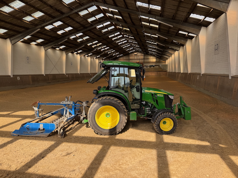 John Deere Kommunaltraktor beim Abziegen der Reitböden in einer Reithalle Dressur