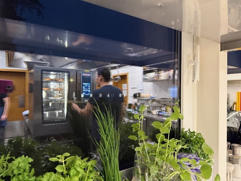 Blick durch das Küchenfenster der Hofküche auf Koch Stefan bei der Arbeit, im Vordergrund frische Kräuter wie Schnittlauch, Petersilie und Minze.