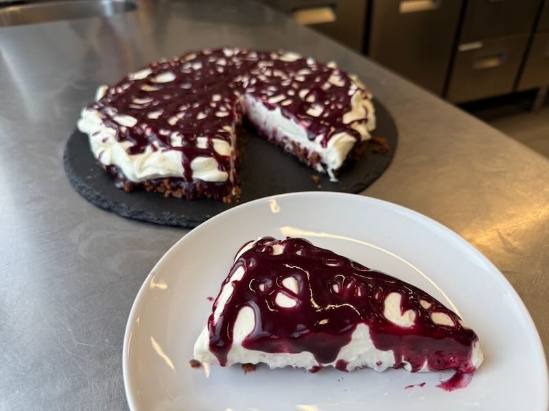 Heidelbeer-Schmand-Torte mit knusprigem Boden und Beerenspiegel, angeschnitten und mit einem Stück auf dem Teller in der Hofküche auf Gut Tangstedt.