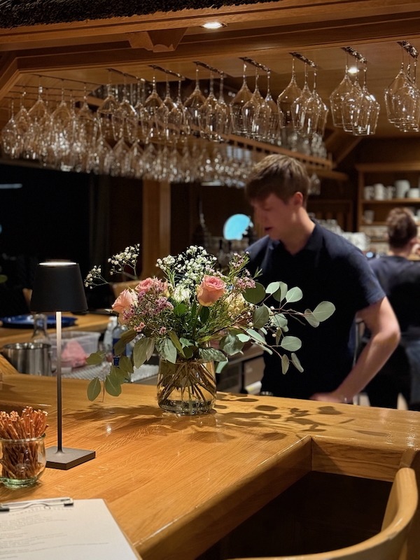 Blumengesteck auf der Theke der Hofküche Gut Tangstedt mit Blick auf das Serviceteam hinter der Bar – gemütliche Atmosphäre für Feiern und Veranstaltungen