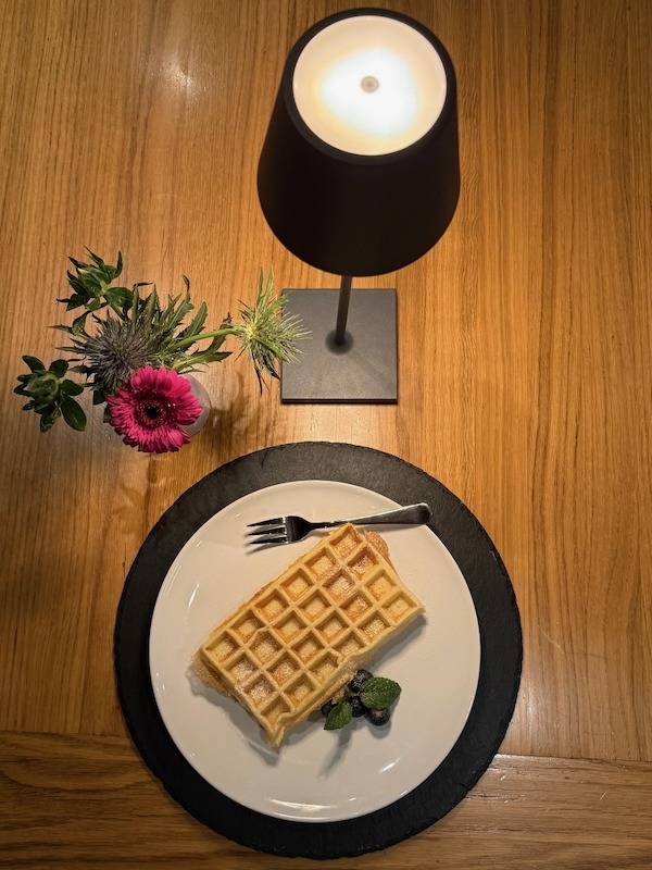 Frisch gebackene belgische Waffel mit Puderzucker, Beeren und Minze, serviert auf Schieferplatte in der Hofküche Gut Tangstedt unter warmem Lampenlicht