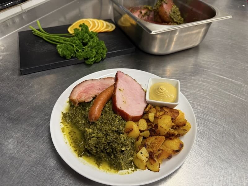 Hausgemachter Grünkohl mit Kassler, Pinkel und Bratkartoffeln, serviert mit Senf auf einem Teller in der Hofküche auf Gut Tangstedt.