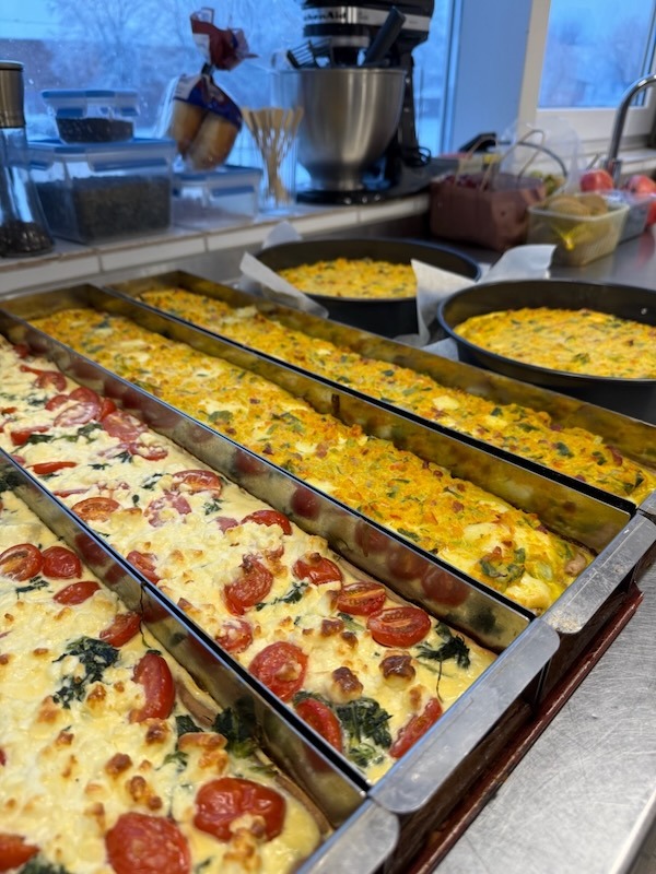 Mehrere Backbleche mit frisch zubereiteten Quiches, darunter vegetarische Varianten mit Tomaten und Spinat in der Profiküche.