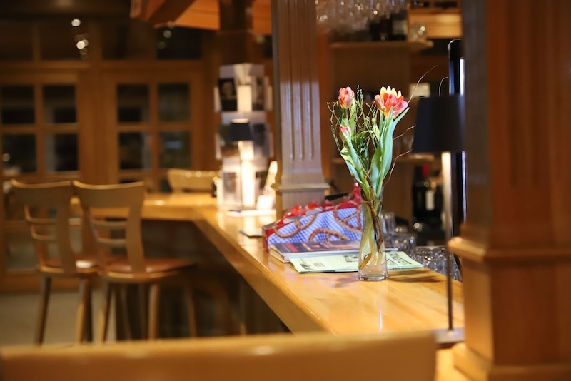 Einladender hölzerner Bartresen im Bistro mit frischen Tulpen in einer Vase und gemütlichen Barhockern im Hintergrund.