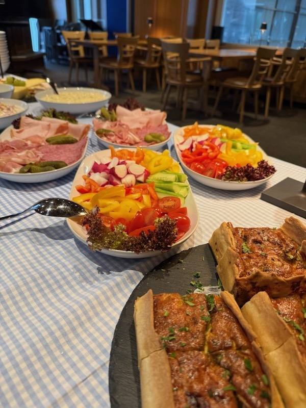 Reichhaltiges Brunch-Buffet mit Platten von Aufschnitt, frischen Gemüsesticks wie Paprika und Tomaten sowie herzhafter Quiche auf einer blau-weiß karierten Tischdecke.