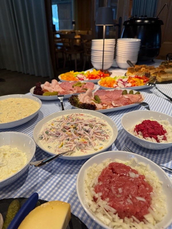 Herzhaftes Frühstücksbuffet mit frischem Mett und Zwiebeln, hausgemachten Salaten, Wurstplatten mit Schinken und Salami sowie Beilagen aus Paprika, Radieschen und Gewürzgurken.