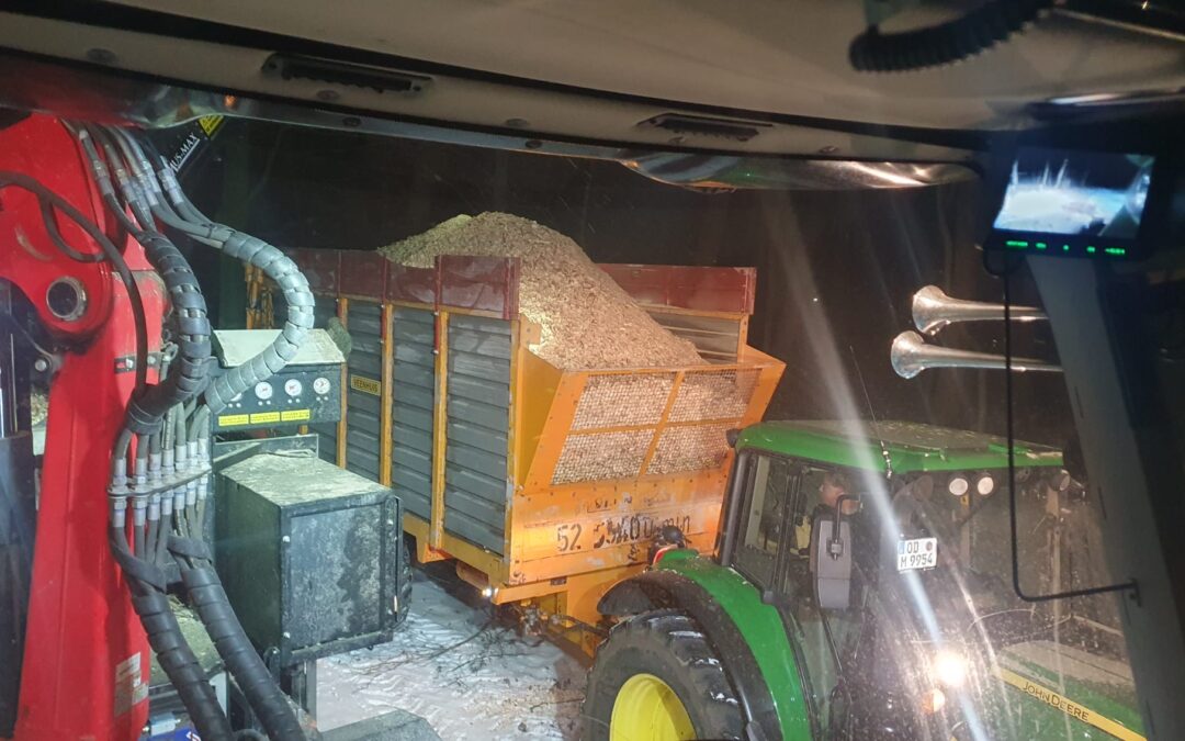 Nächtliche Produktion von Holzhackschnitzeln auf Gut Tangstedt: Ein Traktor mit beladenem Anhänger steht im Scheinwerferlicht neben einer Häckselmaschine im verschneiten Außenbereich.