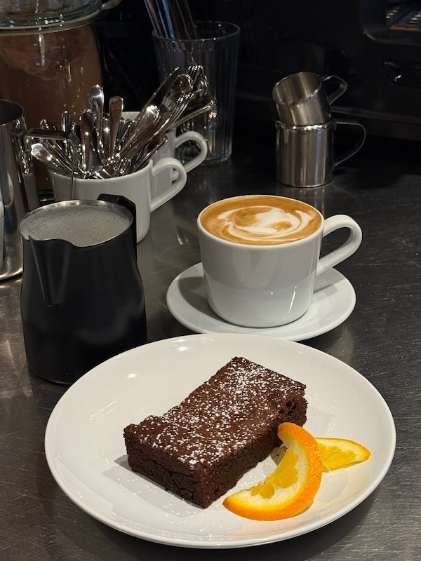 Ein saftiges Stück Schokoladenkuchen mit Puderzucker und Orangengarnitur, serviert mit einer Tasse Cappuccino auf einem Tresen.
