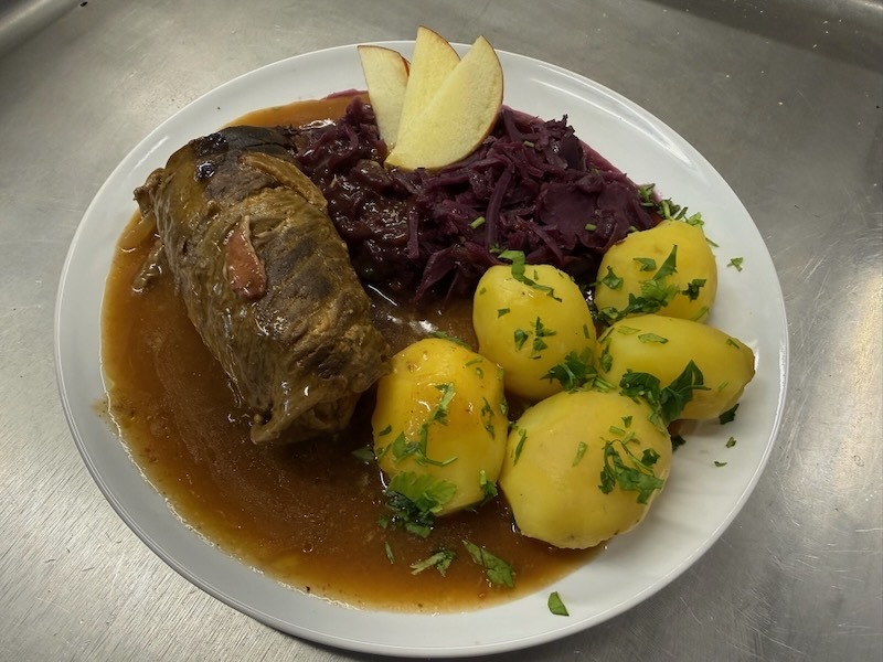 Traditionelle Rinderroulade in dunkler Sauce, serviert mit Apfel-Rotkohl und Petersilienkartoffeln auf einem wei&szlig;en Teller.
