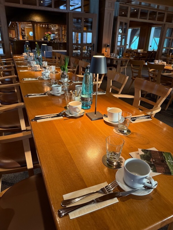 ange, festlich gedeckte Holztafel in einem Restaurant, vorbereitet für ein Frühstücksbuffet mit weißen Kaffeetassen, Besteck und moderner Tischbeleuchtung in gemütlichem Ambiente.