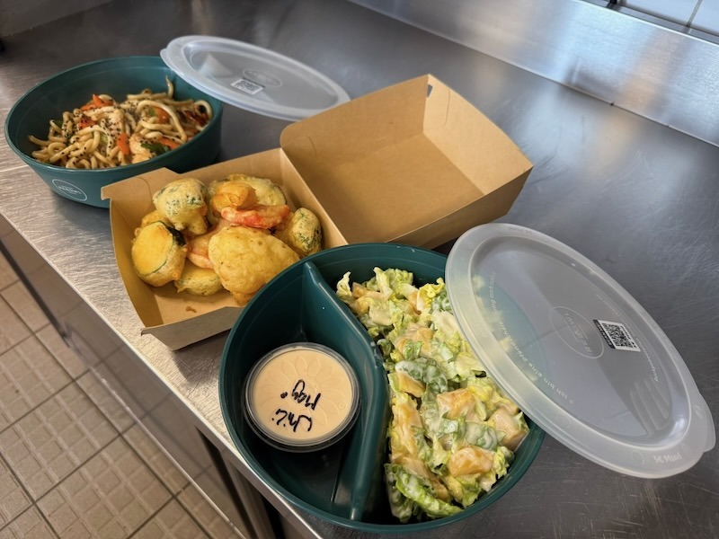 Verschiedene asiatische Gerichte wie Udon-Nudeln, Gemüse-Tempura und frischer Salat, nachhaltig verpackt in Mehrweg-Pfandsystem-Behältern (Rebowl) für den Take-away-Service.