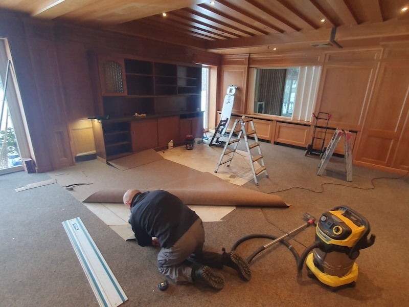 Handwerker bei Bodenverlegearbeiten in einem holzgetäfelten Veranstaltungsraum mit großem Tresen und Renovierungsequipment.