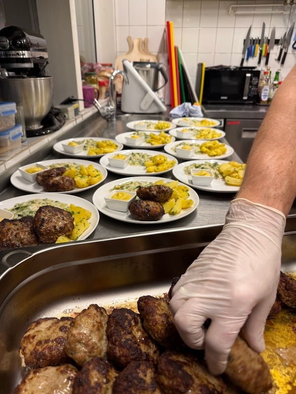 Frisches Anrichten von hausgemachten Frikadellen mit Petersilienkartoffeln und Rahmgemüse auf Tellern in der Hofküche.