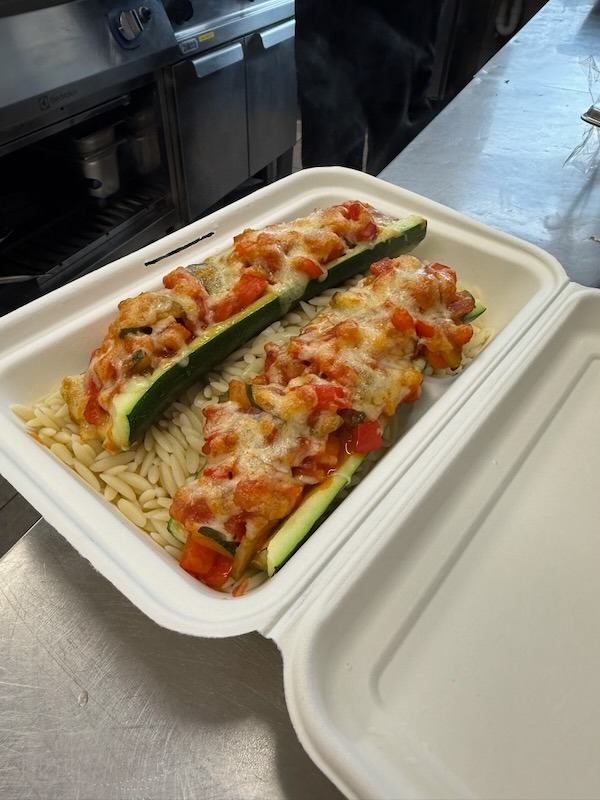 Vegetarisch gefüllte Zucchini mit Käse überbacken auf einem Bett aus Kritharaki-Reisnudeln, serviert in einer Take-away-Box.