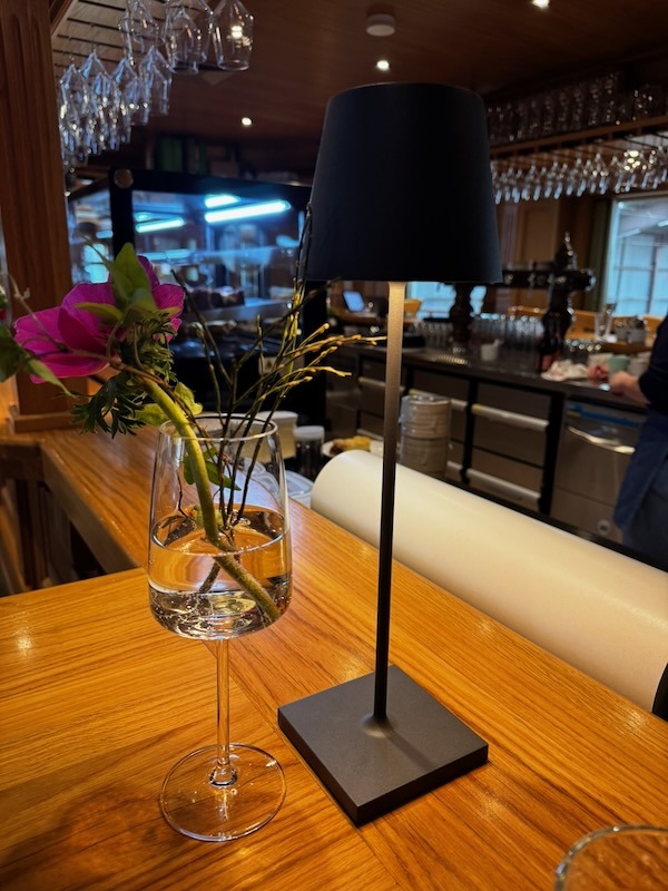 Stimmungsvolles Detail des hölzernen Bartresens in der Hofküche mit einer modernen schwarzen LED-Tischleuchte und einer pinken Blume in einem Weinglas.