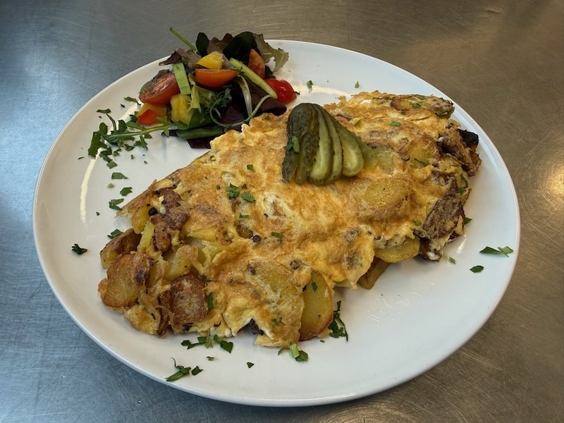 Herzhaftes Bauernfrühstück mit goldbraunen Bratkartoffeln und Ei, garniert mit Gewürzgurken und serviert mit einem frischen Beilagensalat auf einem weißen Teller.