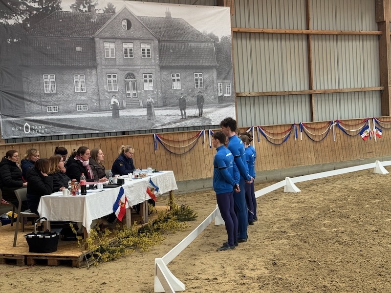 Richtergremium an einem Tisch in der Reithalle während der Feedback-Runde mit einer Voltigiermannschaft vor einer historischen Gutshof-Kulisse.