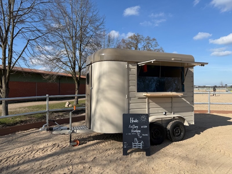 Mobiler Café-Anhänger der Hofküche Gut Tangstedt – ein cremefarbener umgebauter englischer Pferdeanhänger mit aufgeklapptem Verkaufsfenster und Holzablage, aufgestellt auf sandigem Untergrund neben den Roundpens am Beekmorweg. Im Vordergrund eine Kreidetafel mit dem Hinweis auf die Eröffnungsveranstaltung.