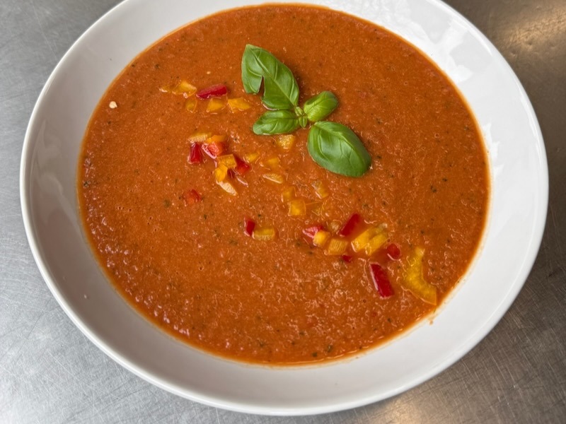 Leuchtend rote Gazpacho in einem tiefen wei&szlig;en Teller, garniert mit fein gew&uuml;rfelter roter und gelber Paprika sowie frischen Basilikumbl&auml;ttern.