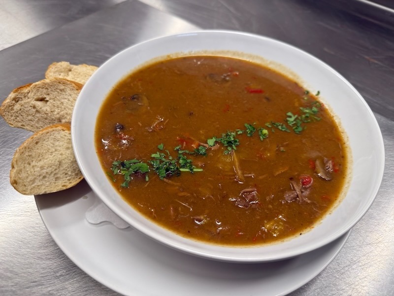 ampfende Gulaschsuppe mit reichlich Fleischeinlage und Pilzen, serviert mit drei Scheiben frischem Baguette.