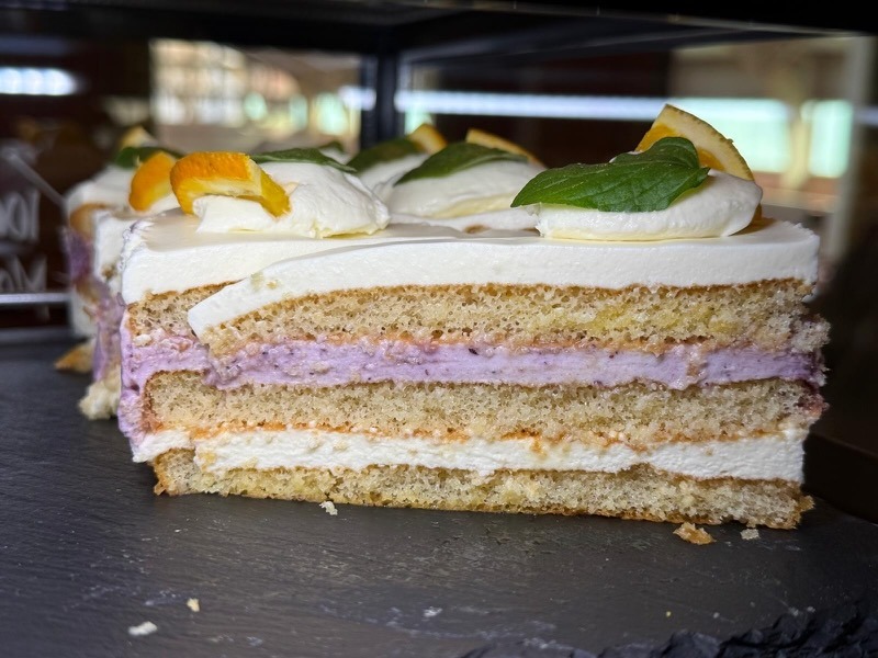 Querschnitt einer mehrstöckigen Käse-Sahnetorte mit hellen Bisquitböden, einer lila Beerencrème- und einer orangefarbenen Fruchtschicht, weißer Sahnecreme sowie Dekoration aus Orangenscheiben und frischer Minze.