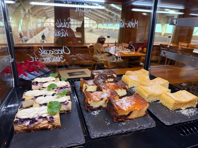 Kuchentheke der Hofküche Gut Tangstedt mit täglich frisch gebackenen Kuchen. Im Hintergrund Tische der Gaststube und die Reithalle, in der schemenhaft ein Reiter zu erkennen ist.