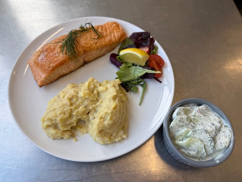 Gebratenes Lachsfilet mit Dillgarnitur und Zitronenscheibe auf einem wei&szlig;en Teller, serviert mit cremigem Kartoffelp&uuml;ree und einem kleinen Gurkensalat.