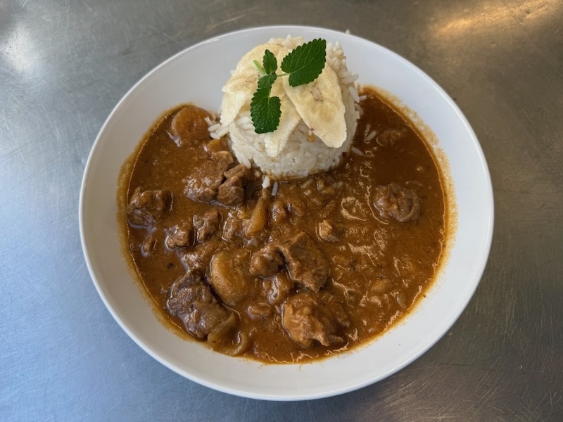 Afrikanisches Lammragout in w&uuml;rziger, dunkelbrauner Sauce mit Pilzen, serviert in einem tiefen wei&szlig;en Teller. In der Mitte ein gest&uuml;rzter Reistimbale, garniert mit Bananenchips und frischer Minze.