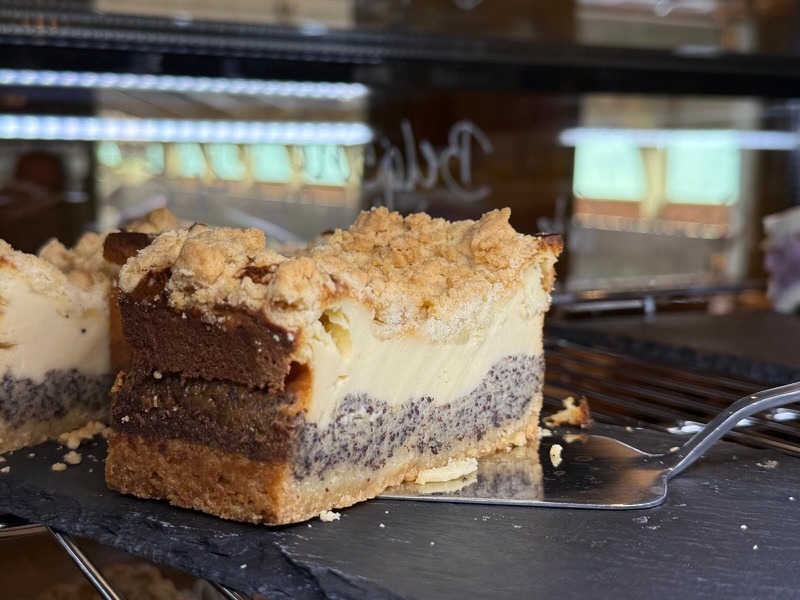 Aufgeschnittenes Stück Mohn-Käsekuchen mit sichtbaren Schichten: Mürbeteigboden, Mohnfüllung, cremige Käsemasse und goldbraunen Streuseln obenauf, präsentiert auf einer Schieferplatte in einer Kuchen-Theke.
