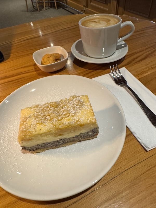 Ein Stück saftiger, hausgebackener Mohn-Käsekuchen mit Streuseln und Puderzucker, serviert mit einer Tasse Cappuccino und feinem Gebäck auf einem Holztisch.