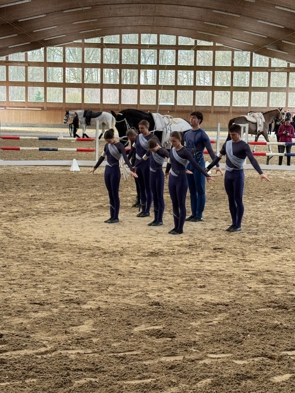 Eine Voltigiermannschaft in einheitlichen blau-grauen Trikots präsentiert sich aufgestellt in der Mitte einer großen Reithalle.