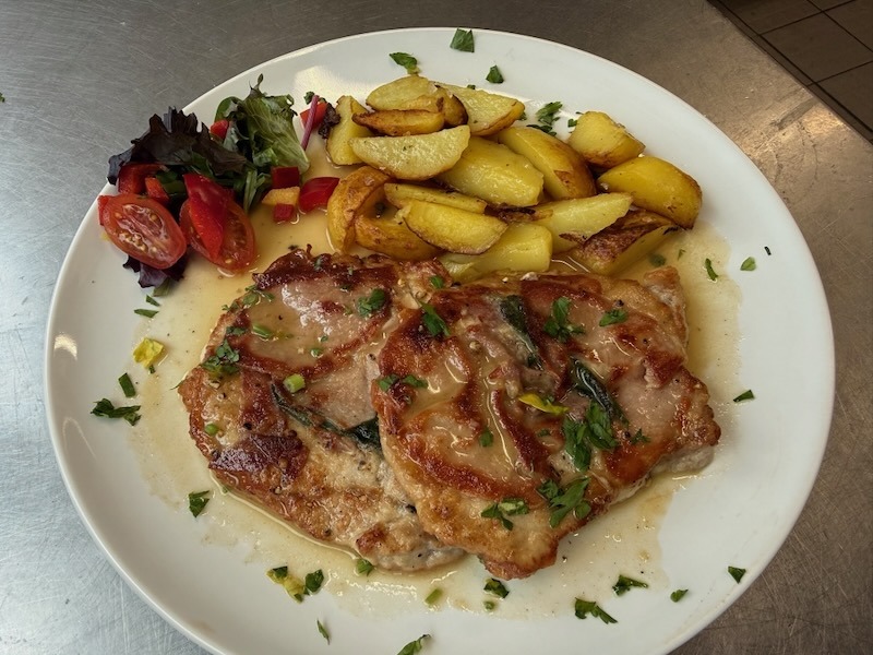 Zwei zart gebratene Fleischst&uuml;cke nach Saltimbocca-Art mit frischem Salbei und Schinken, serviert mit goldbraunen Bratkartoffeln und einem kleinen Beilagensalat auf einem wei&szlig;en Teller.