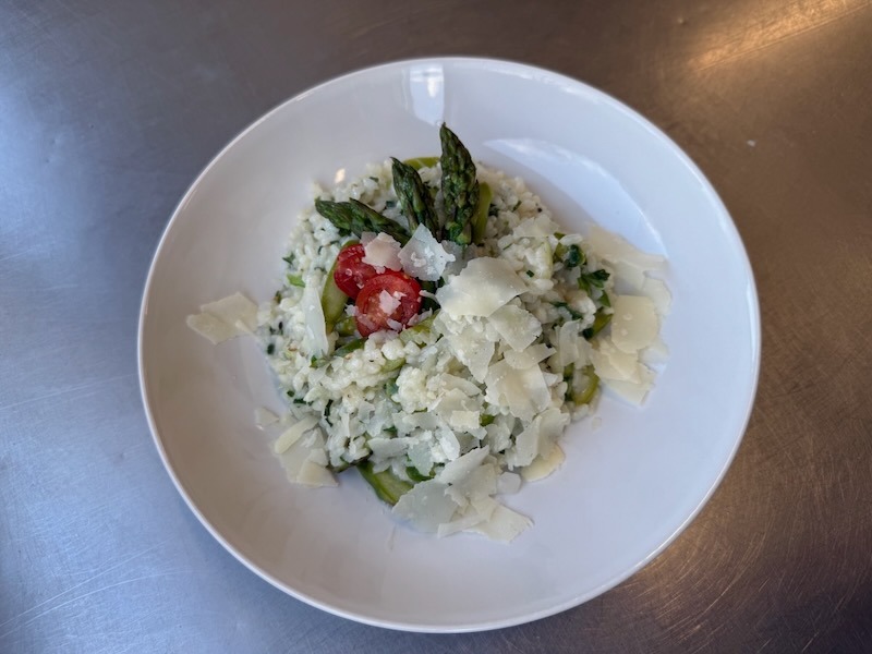 Cremiges Spargelrisotto mit gr&uuml;nem Spargel auf einem wei&szlig;en Teller, garniert mit Kirschtomaten und gro&szlig;z&uuml;gig gehobeltem Parmesan.