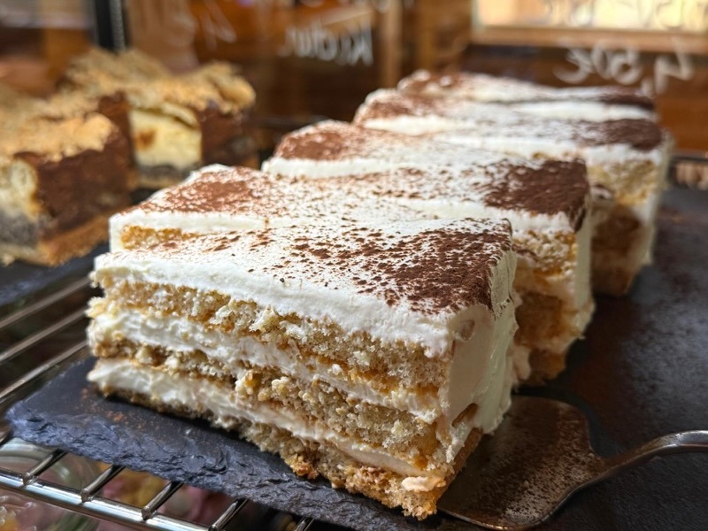 Mehrere Stücke Tiramisu-Torte mit abwechselnden Schichten aus Löffelbiskuit und Mascarpone-Creme, großzügig mit Kakaopulver bestäubt, auf einer Schieferplatte angerichtet.