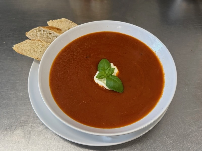 Herzhafte Tomatensuppe mit einer cremigen Schmandhaube und frischem Basilikum, serviert mit knusprigen Baguettescheiben auf einem weißen Teller.