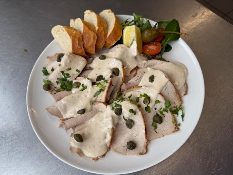 Vitello Tonnato mit zart rosa gegartem Schweinefleisch, cremiger Thunfischsauce und Kapern, serviert mit Baguettescheiben und einer frischen Salatbeilage.