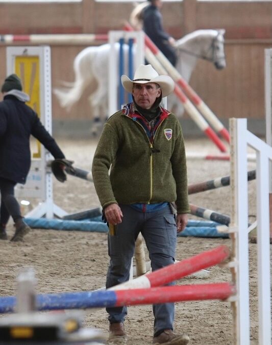 Trainer Sven Vogelsang mit weißem Cowboyhut und grüner Fleecejacke läuft zwischen Springstangen in der Reithalle von Gut Tangstedt, im Hintergrund ein weißes Pferd mit Reiter.