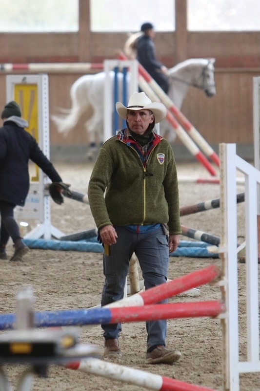 Trainer Sven Vogelsang mit weißem Cowboyhut und grüner Fleecejacke läuft zwischen Springstangen in der Reithalle von Gut Tangstedt, im Hintergrund ein weißes Pferd mit Reiter.