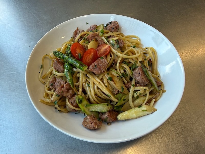 Linguine mit grünem Spargel, weißem Spargel und gebratenen Salsiccia-Stücken in einem weißen tiefen Teller, garniert mit halbierter Kirschtomate und frisch gehackten Kräutern.