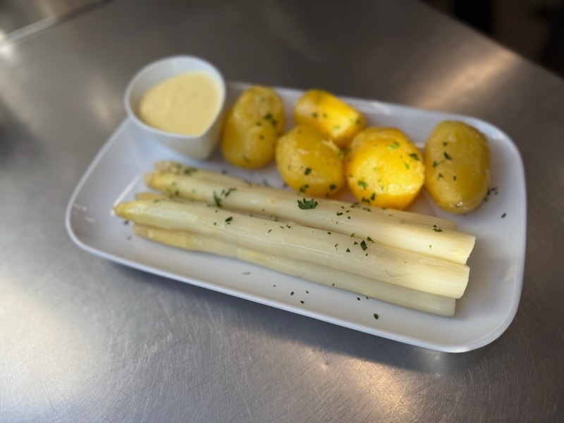 Klassisches Spargelgericht auf einem weißen Servierteller: drei Stangen weißer Spargel mit Petersilie bestreut, dazu butterglasierte Salzkartoffeln und ein Schälchen Sauce Hollandaise.