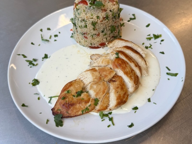 Aufgeschnittene gebratene Hähnchenbrust auf cremiger Joghurtsoße mit frischer Petersilie, angerichtet neben einem gestürzten Timbale aus Couscous-Tabulé mit Tomaten und Kräutern.