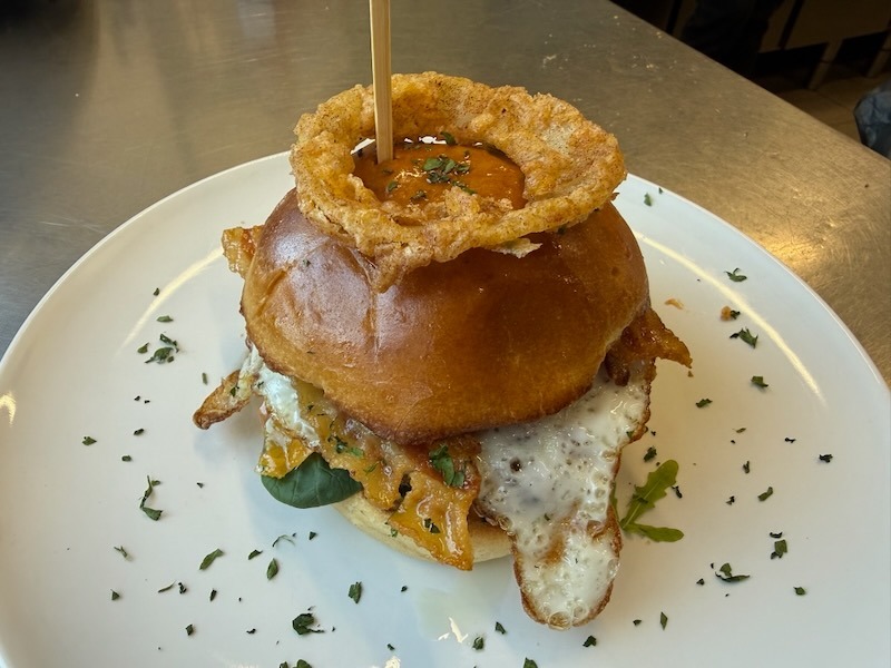 Üppig belegter Bacon-Burger auf goldenem Brioche-Bun mit Spiegelei, Cheddar, Bacon und frischem Blattgemüse, gekrönt von einem knusprigen Onion Ring mit BBQ-Sauce, fixiert mit einem Holzspieß.