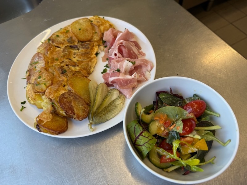 Bauernfrühstück aus Bratkartoffeln mit Rührei und frischen Kräutern auf einem weißen ovalen Teller, dazu rosa Kochschinken und Gewürzgurken. Als Beilage eine Schüssel bunter Blattsalat mit Kirschtomaten, Gurke und Paprika.
