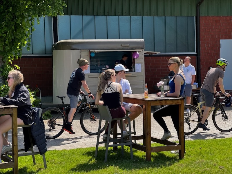 Sommerlicher Pop-up-Biergarten auf dem Gutshof: Ein mobiler Feldküchenwagen steht neben einem grün verkleideten Backsteingebäude, Gäste stehen an und sitzen im Hintergrund unter großen Bäumen bei blauem Himmel.