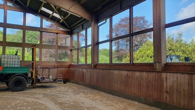 Innenansicht einer Reithalle mit großer Glasfront und Holzverkleidung während der Frühjahrsreinigung. Ein Traktor mit Anhänger und Reinigungsgerät steht im sandbedeckten Hallenbereich, im Hintergrund grüne Bäume bei blauem Himmel.