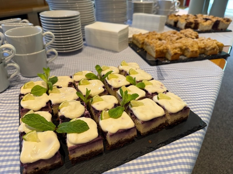 Kuchenbuffet auf einem mit blau-weißem Stoff gedeckten Tisch: Im Vordergrund auf einer Schieferplatte gleichmäßig angerichtete Blaubeer-Käsekuchen-Schnitten mit weißer Creme, Limettenspalten und frischer Minze. Im Hintergrund eine weitere Schieferplatte mit Streuselkuchen, daneben gestapelte weiße Kaffeetassen und Teller.